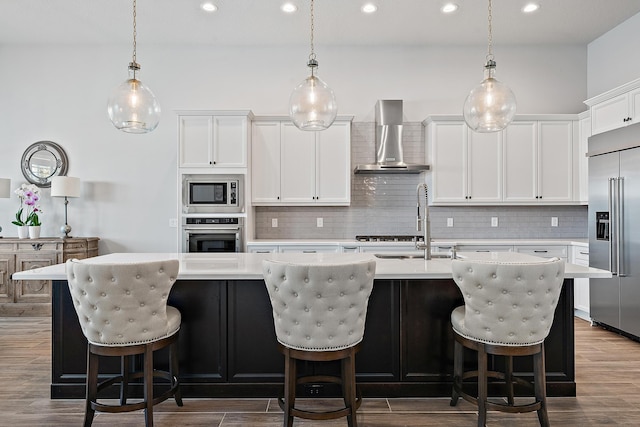 kitchen with built in appliances, a center island with sink, light countertops, and wall chimney exhaust hood