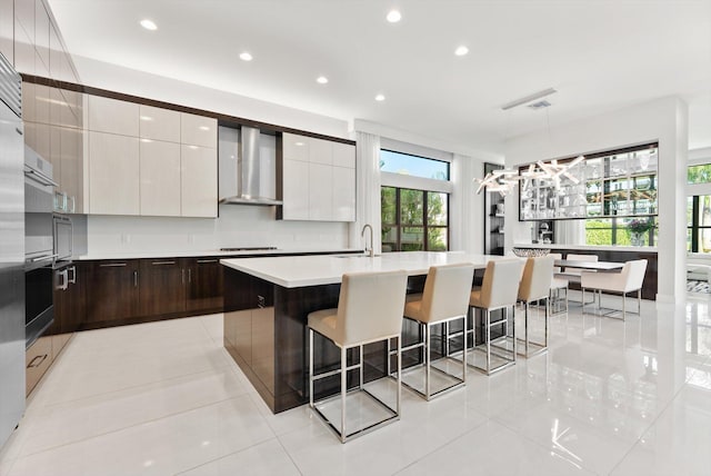 kitchen with a breakfast bar, light countertops, a kitchen island with sink, modern cabinets, and wall chimney exhaust hood