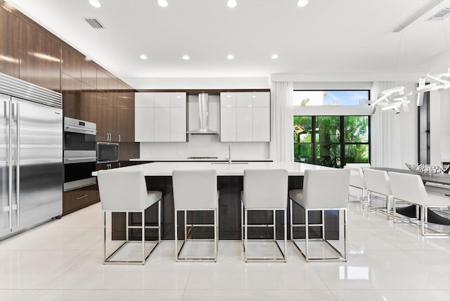 kitchen with wall chimney range hood, modern cabinets, a breakfast bar, and built in appliances