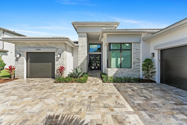 entrance to property with stone siding, an attached garage, decorative driveway, french doors, and stucco siding