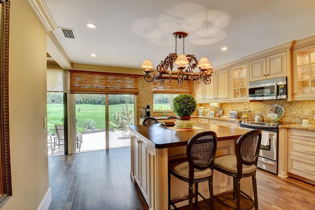 kitchen with wood finished floors, appliances with stainless steel finishes, cream cabinetry, tasteful backsplash, and glass insert cabinets