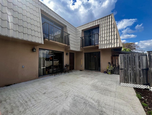 rear view of property with a patio, stucco siding, mansard roof, fence, and a balcony