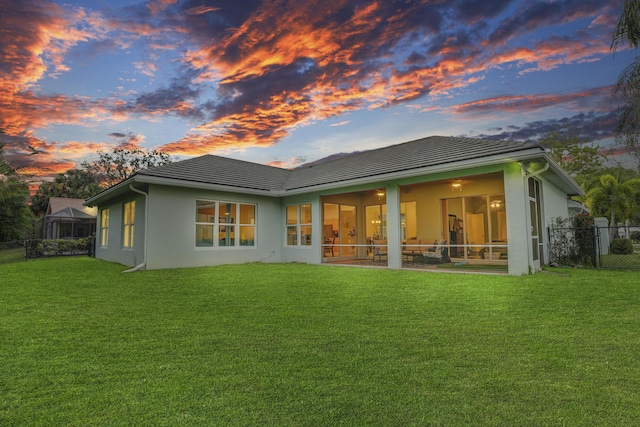 back of property at dusk featuring a lawn, a sunroom, a tiled roof, fence, and stucco siding