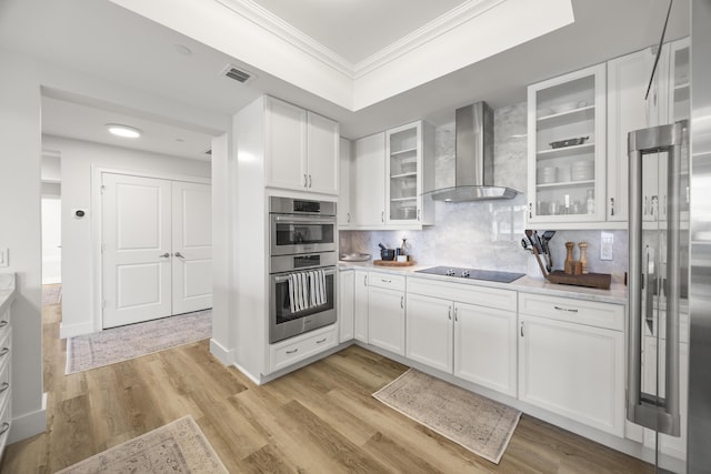 kitchen with crown molding, stainless steel appliances, white cabinets, wall chimney range hood, and light wood-type flooring