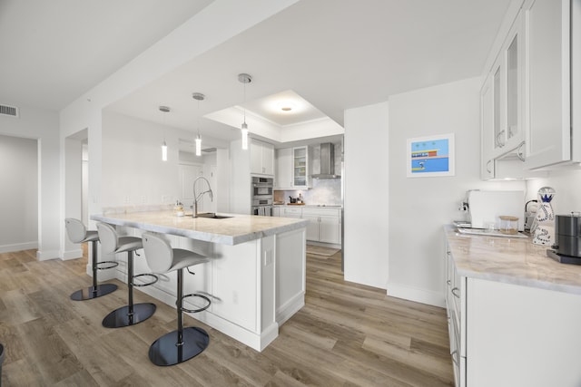 kitchen featuring a kitchen breakfast bar, a tray ceiling, wall chimney range hood, white cabinetry, and a sink