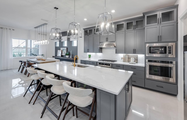 kitchen featuring gray cabinetry, under cabinet range hood, a sink, appliances with stainless steel finishes, and a center island with sink