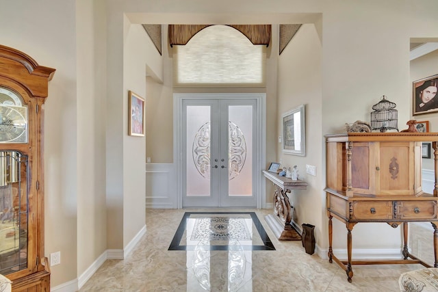 entryway featuring french doors, marble finish floor, a decorative wall, a towering ceiling, and baseboards