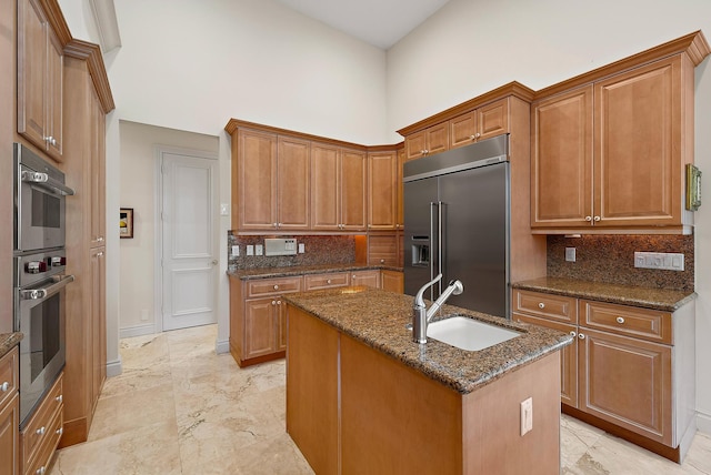 kitchen featuring stainless steel appliances, a sink, marble finish floor, brown cabinets, and dark stone countertops