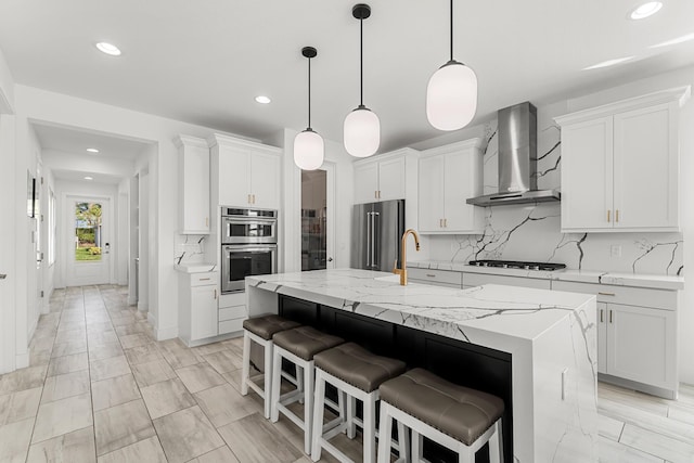 kitchen with decorative backsplash, appliances with stainless steel finishes, a kitchen island with sink, a sink, and wall chimney exhaust hood