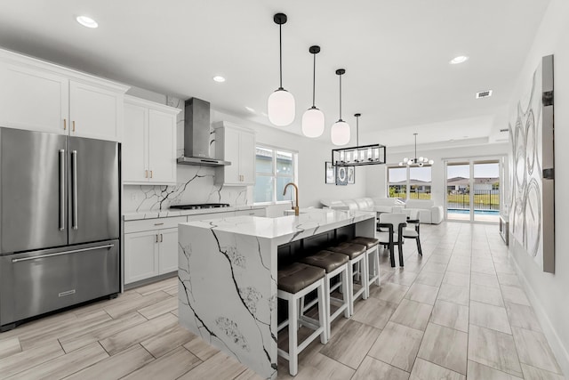 kitchen with high end fridge, backsplash, white cabinets, stovetop, and wall chimney exhaust hood