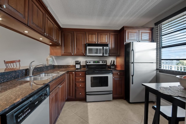 kitchen with a sink, a textured ceiling, dark stone counters, appliances with stainless steel finishes, and light tile patterned flooring