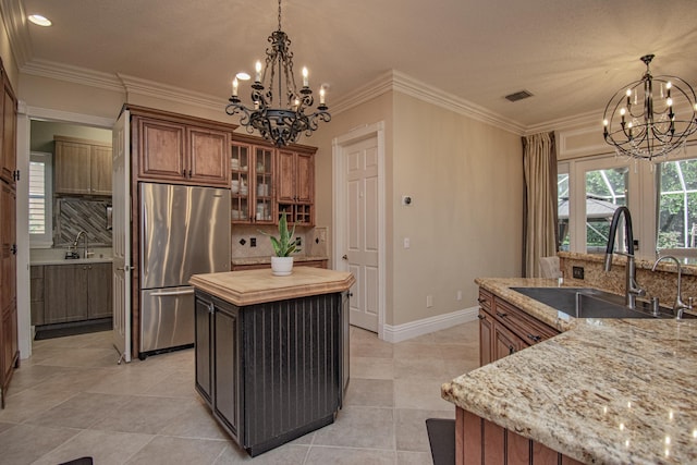 kitchen featuring an inviting chandelier, brown cabinetry, a kitchen island with sink, a sink, and stainless steel fridge