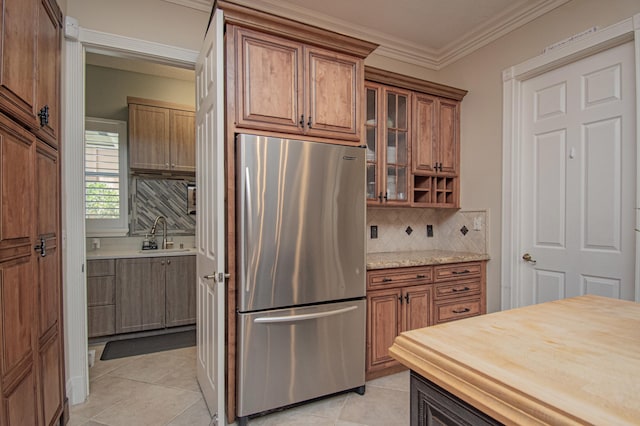 kitchen featuring freestanding refrigerator, light tile patterned floors, brown cabinetry, ornamental molding, and a sink