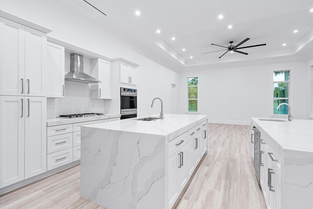 kitchen with a raised ceiling, wall chimney range hood, an island with sink, and a sink