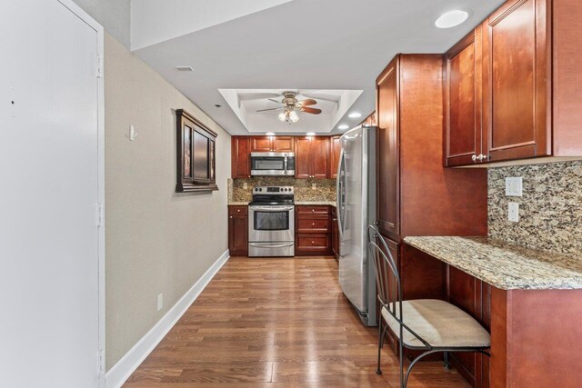 kitchen with baseboards, appliances with stainless steel finishes, wood finished floors, a raised ceiling, and a ceiling fan