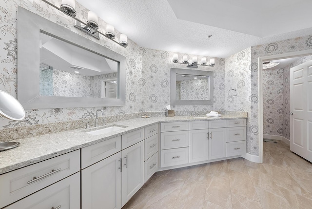 bathroom with a textured ceiling, double vanity, a sink, and wallpapered walls