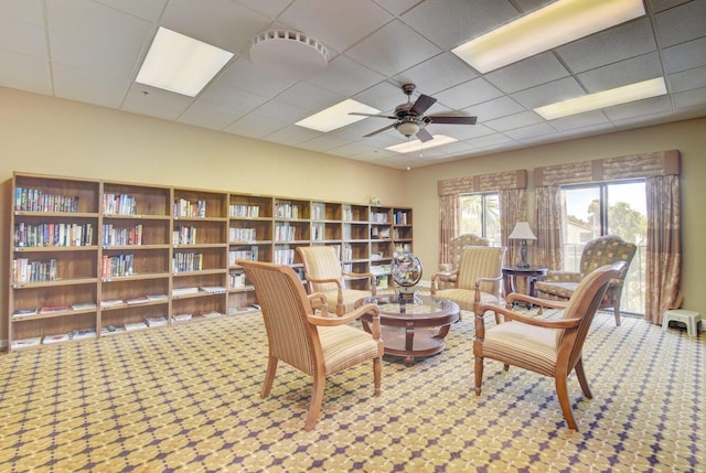 living area with ceiling fan, bookshelves, a drop ceiling, and carpet flooring