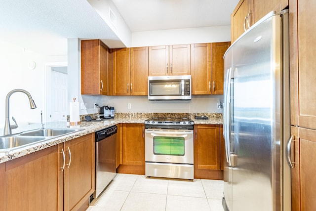 kitchen featuring light stone counters, light tile patterned floors, brown cabinetry, a sink, and appliances with stainless steel finishes