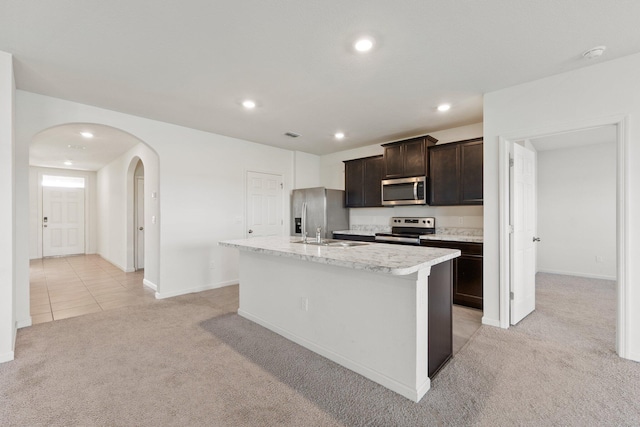 kitchen with a center island with sink, arched walkways, light colored carpet, stainless steel appliances, and dark brown cabinets