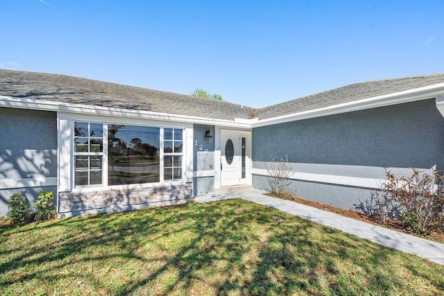 view of exterior entry featuring roof with shingles, a lawn, and stucco siding