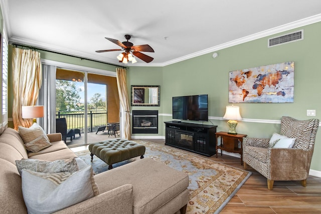 living area featuring visible vents, a glass covered fireplace, ceiling fan, ornamental molding, and wood finished floors