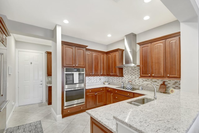 kitchen with double oven, light stone counters, a peninsula, a sink, and wall chimney range hood