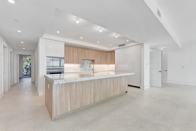 kitchen with tasteful backsplash, a raised ceiling, modern cabinets, a large island with sink, and light brown cabinets