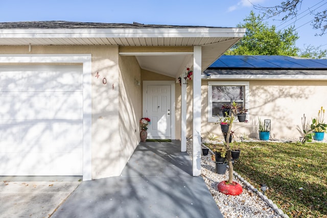 view of exterior entry featuring a garage, solar panels, a shingled roof, and stucco siding