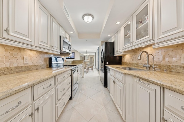 kitchen featuring light tile patterned floors, a sink, appliances with stainless steel finishes, a tray ceiling, and glass insert cabinets