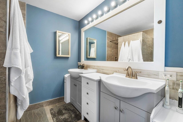 full bathroom featuring baseboards, two vanities, a sink, and tiled shower