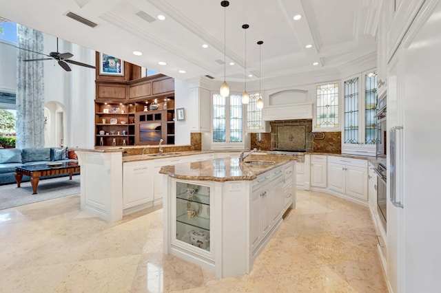 kitchen with glass insert cabinets, an island with sink, ornamental molding, and a ceiling fan