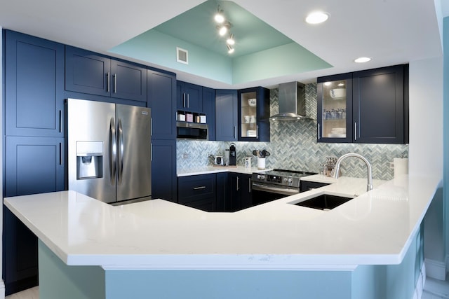 kitchen featuring a peninsula, a tray ceiling, stainless steel appliances, wall chimney range hood, and a sink