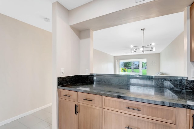 kitchen with baseboards, light brown cabinetry, dark stone counters, light tile patterned floors, and a notable chandelier