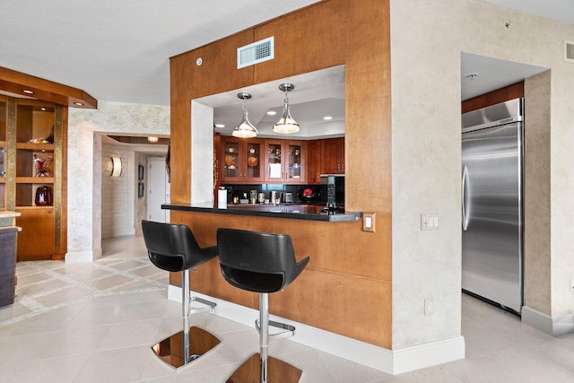 kitchen with light tile patterned floors, visible vents, dark countertops, glass insert cabinets, and stainless steel built in fridge