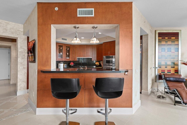 kitchen featuring a breakfast bar, dark countertops, visible vents, brown cabinetry, and glass insert cabinets