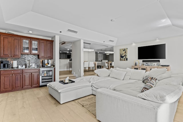 living area featuring wine cooler, visible vents, light wood-style floors, indoor wet bar, and a raised ceiling