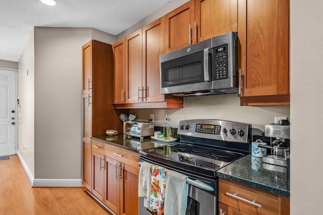 kitchen featuring stainless steel appliances, light wood-style flooring, brown cabinetry, dark stone counters, and baseboards