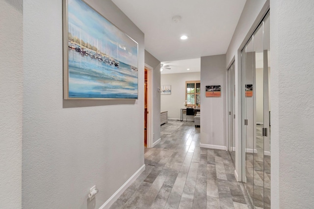 hallway featuring wood finished floors, a textured wall, baseboards, and recessed lighting