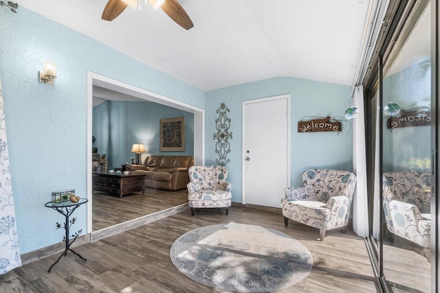 living area featuring a ceiling fan, a textured wall, vaulted ceiling, and wood finished floors