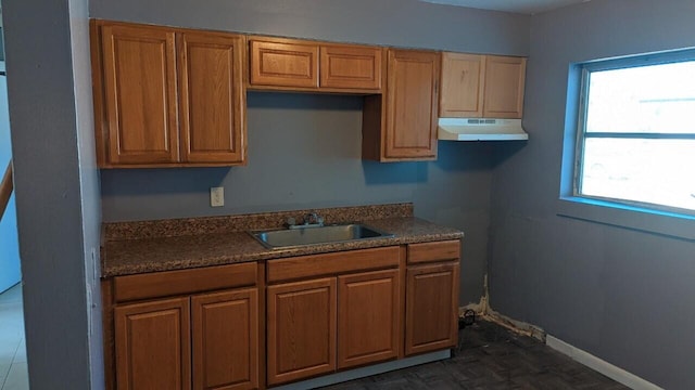 kitchen featuring brown cabinets, dark countertops, a sink, under cabinet range hood, and baseboards