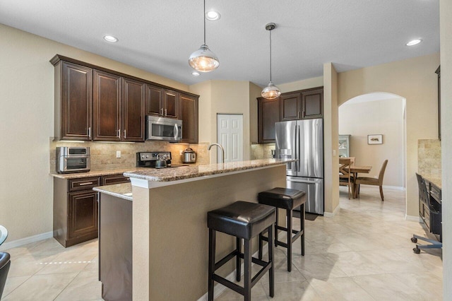 kitchen featuring dark brown cabinetry, tasteful backsplash, arched walkways, appliances with stainless steel finishes, and a kitchen bar