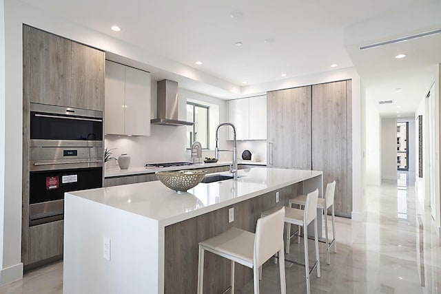 kitchen with modern cabinets, a breakfast bar, a sink, double oven, and wall chimney exhaust hood