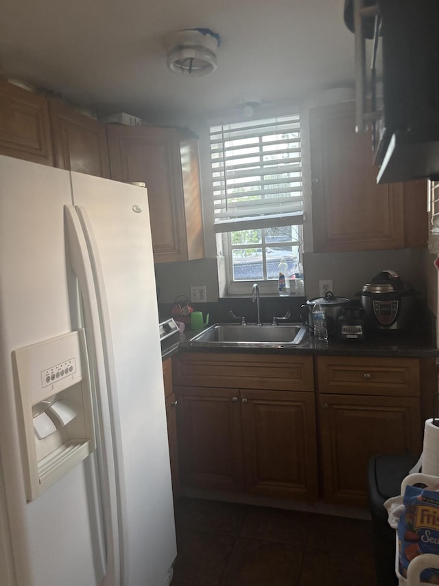 kitchen with brown cabinets, dark countertops, a sink, white fridge with ice dispenser, and dark tile patterned floors