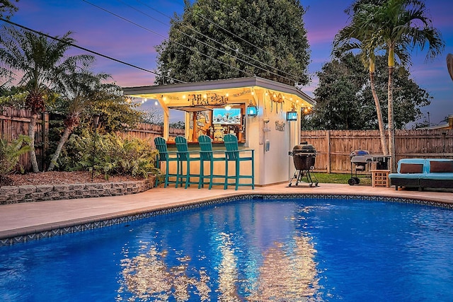 pool at dusk with a fenced in pool, outdoor dry bar, area for grilling, a patio area, and a fenced backyard