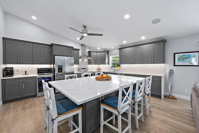 kitchen with beverage cooler, wall chimney exhaust hood, a kitchen island, appliances with stainless steel finishes, and vaulted ceiling