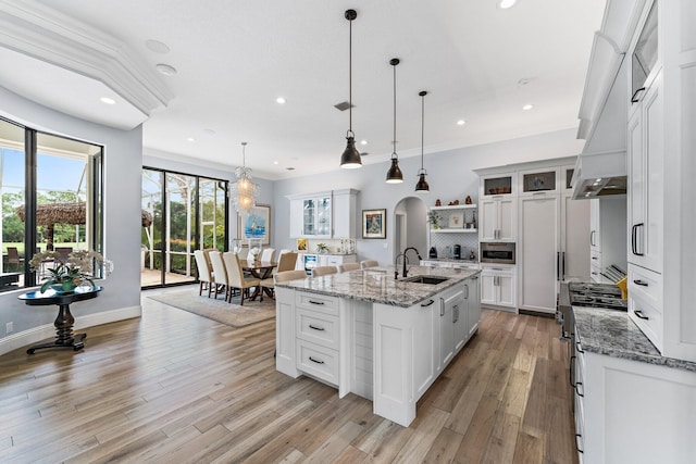 kitchen with high end stove, an island with sink, arched walkways, white cabinetry, and a sink