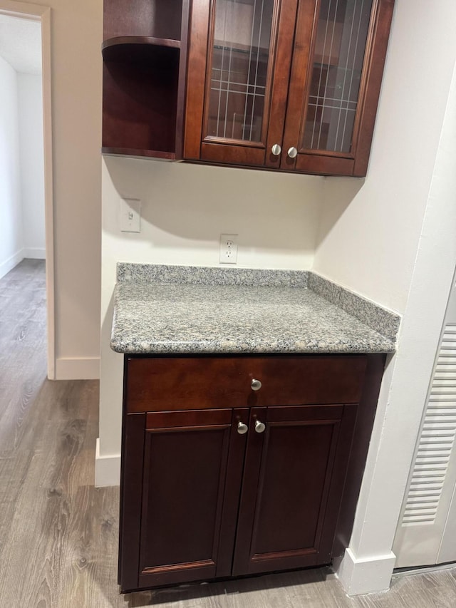 kitchen with light wood finished floors, glass insert cabinets, dark brown cabinets, light stone countertops, and baseboards