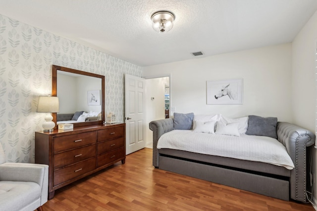 bedroom with wood finished floors, visible vents, a textured ceiling, and wallpapered walls