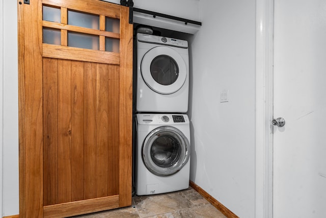 washroom featuring stacked washer / dryer, stone finish flooring, baseboards, and a barn door