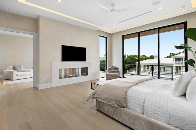 bedroom with access to outside, a glass covered fireplace, a tray ceiling, and wood finished floors